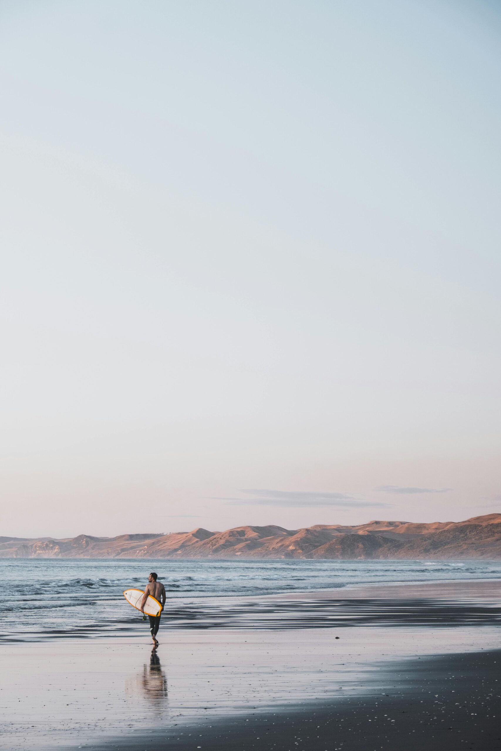 A lone surfer with a surfboard walks along the serene beach at sunset in New Zealand.